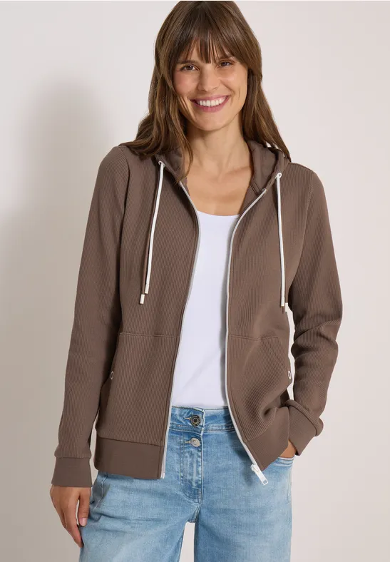 Een lachende vrouw in een bruine hoodie, wit topje en spijkerbroek, voor een neutrale achtergrond.