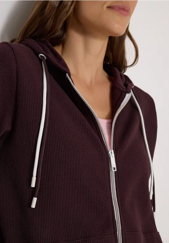 Vrouw draagt een pruimkleurige capuchontrui met ritssluiting en witte koorden over een licht shirt.