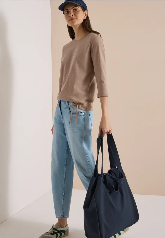 Frau in lässigem Outfit mit beiger Bluse, hellblauen Jeans und Cap hält eine große dunkelblaue Tasche.