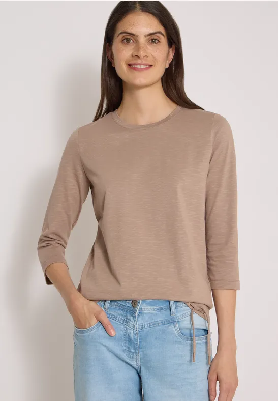 Frau lächelt in beigem 3/4-Arm-Pullover mit Seitenbindung und blauen Jeans vor hellem Hintergrund.