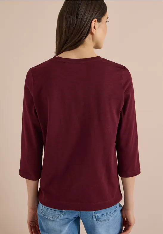 Achteraanzicht van een persoon in een bordeauxrood driekwartmouwshirt en blauwe jeans voor een effen achtergrond.