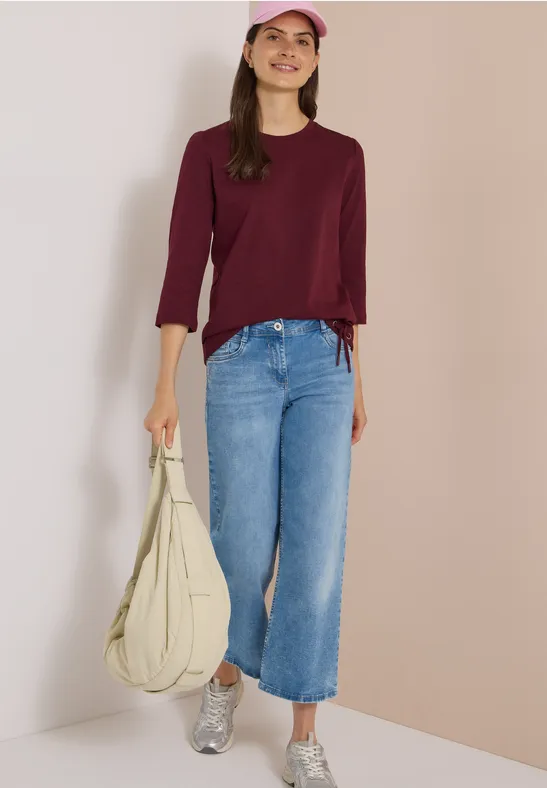Vrouw in bordeaux trui, lichtblauwe jeans, zilveren sneakers, roze pet en grote beige tas.