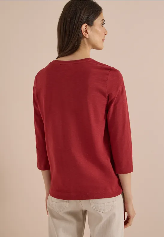 Vrouw rug-aanzicht, draagt rood shirt met driekwartmouw en beige broek.