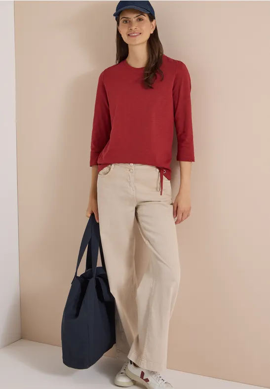 Persoon met rood shirt, beige broek, blauw petje en blauwe tas, staand voor een neutrale achtergrond.