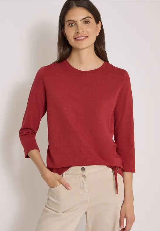 Vrouw in rode driekwart mouwtrui en beige chino, glimlachend met handen in de zakken.