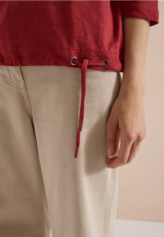Close-up van een persoon met een rood shirt, beige broek en een loshangende koord.