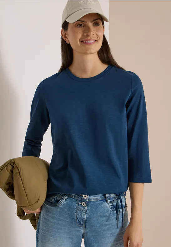 Lächelnde Frau mit Cap, blauem Dreiviertelshirt und Jeans, hält locker eine Jacke.