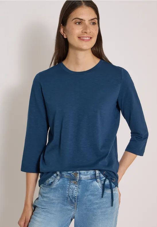 Frau trägt marineblaues 3/4-Arm-Shirt mit seitlicher Schleife und helle Jeans, lächelnd vor hellem Hintergrund.