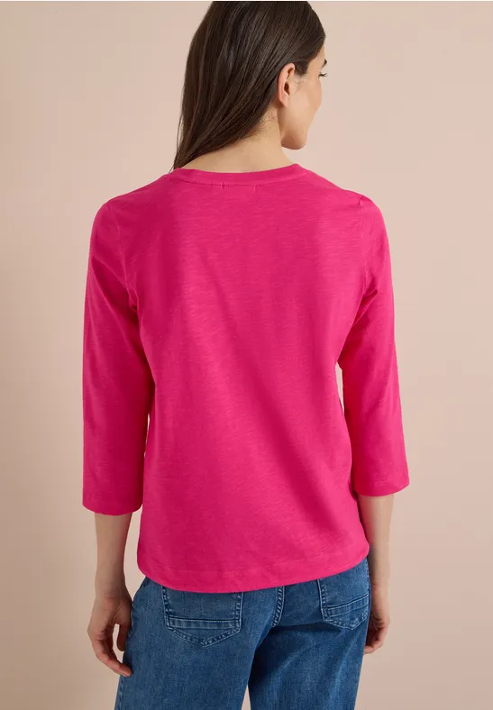 Frau von hinten in pinkem 3/4-Arm-Shirt und blauen Jeans vor hellem Hintergrund.