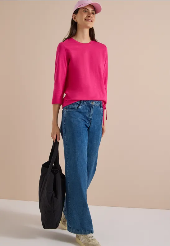 Frau in pinkem Top, weiter blauer Jeans, pinker Cap und schwarzer Tasche vor neutralem Hintergrund.