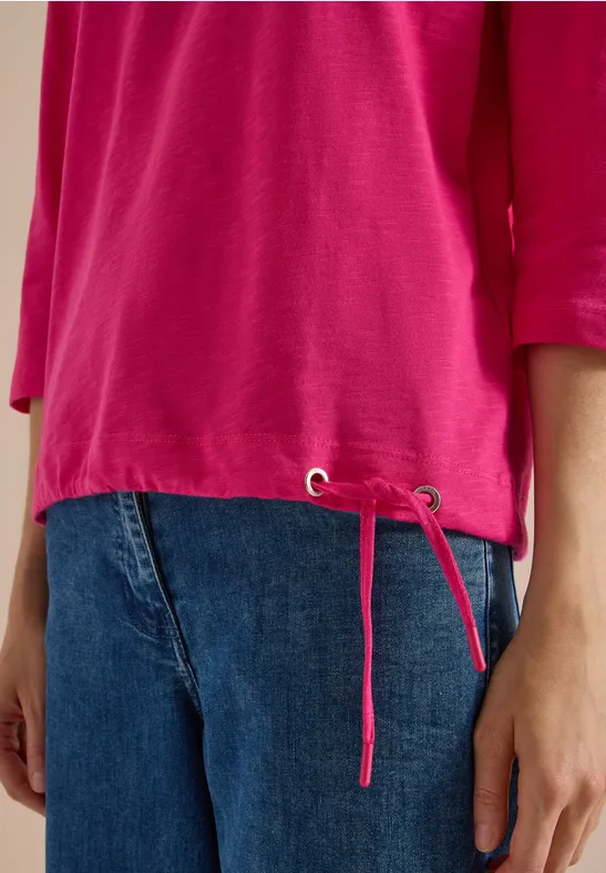 Eine Person trägt ein pinkes Shirt mit seitlichen Kordelzügen und blaue Jeans.