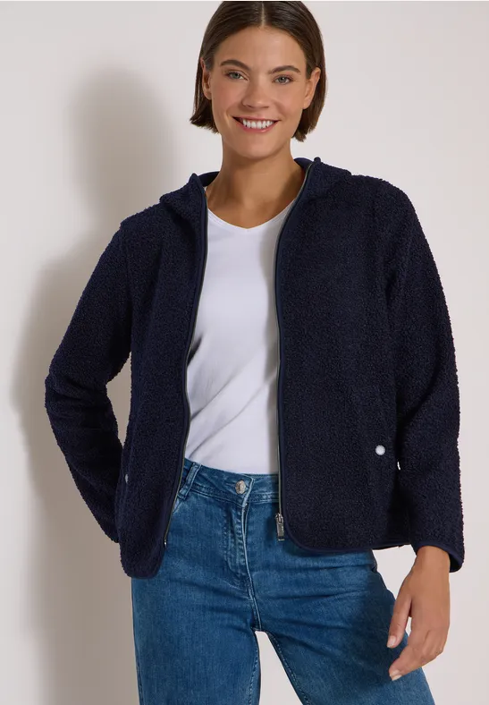 Lächelnde Frau in dunkelblauer Fleecejacke, weißem T-Shirt und Jeans vor hellem Hintergrund.