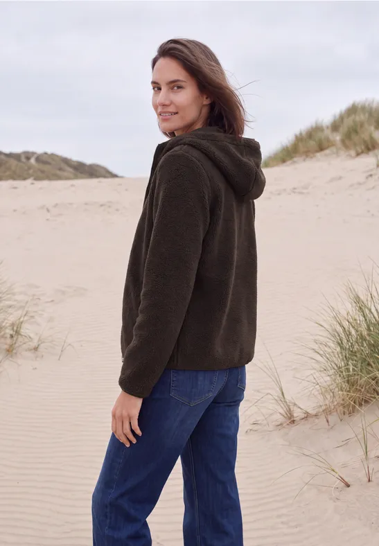Vrouw in bruine capuchonvest en blauwe broek staat op een zandduin en kijkt over haar schouder.
