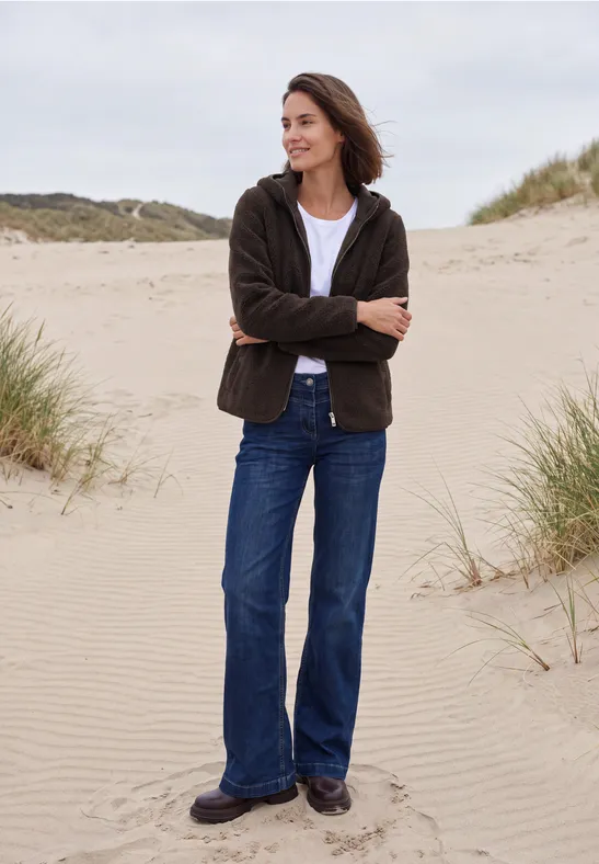 Vrouw in jeans en jas staat ontspannen met gekruiste armen op duinen bij het strand.