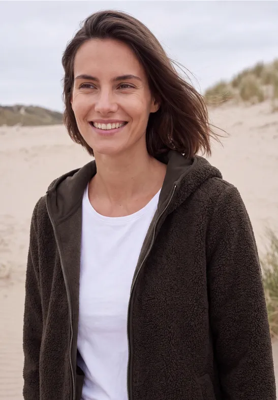 Een glimlachende vrouw in een fleece trui staat op een strand met duinen op de achtergrond.