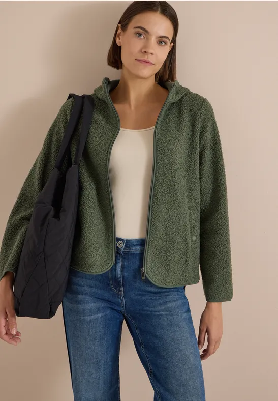 Frau in grüner Fleecejacke, weißem Top, Jeans und schwarzer gesteppter Tasche vor beigem Hintergrund.