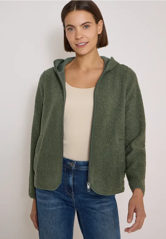 Frau trägt grüne Fleecejacke mit Kapuze, beiges Oberteil und blaue Jeans, vor hellen Hintergrund.
