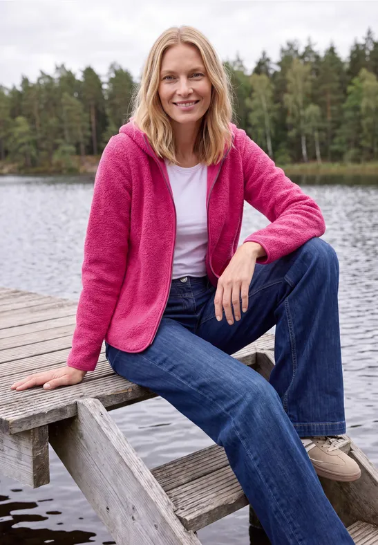 Une femme en veste rose et t-shirt blanc est assise sur une jetée au bord d’un lac.