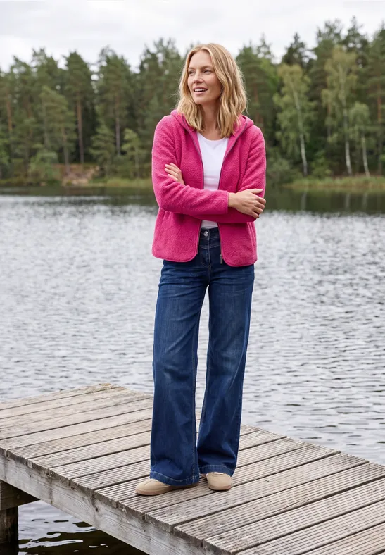 Une femme en pull rose et jean se tient détendue sur un ponton en bois au bord d’un lac, forêt en arrière-plan.