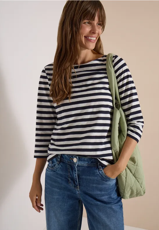 Vrouw in gestreept 3/4-mouwshirt, spijkerbroek en groene gewatteerde schoudertas lacht voor neutrale achtergrond.