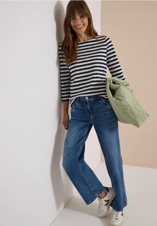 Een glimlachende vrouw in gestreept shirt, blauwe jeans en witte sneakers houdt een groene gewatteerde jas tegen een lichte achtergrond.