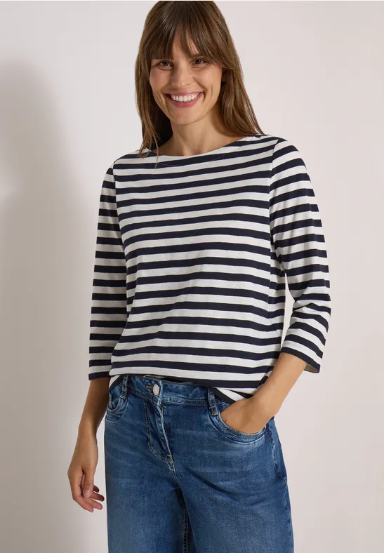 Jonge vrouw lacht in blauw-wit gestreept driekwartshirt en blauwe spijkerbroek tegen effen achtergrond.