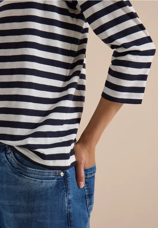 Bovenlijf in blauw-wit gestreept shirt, hand in spijkerbroekzak, voor een beige achtergrond.