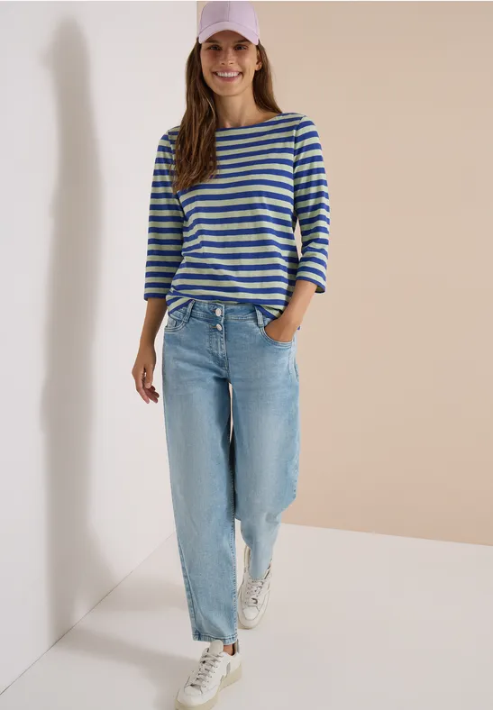 Een lachende vrouw in gestreept shirt, wijde jeans, witte sneakers en pet, staand voor neutrale achtergrond.