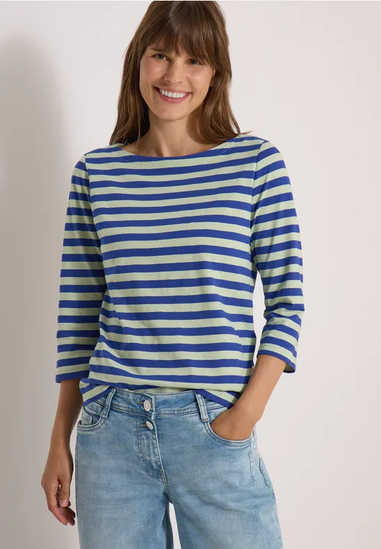 Een glimlachende vrouw draagt een blauw-wit gestreept shirt en een spijkerbroek.