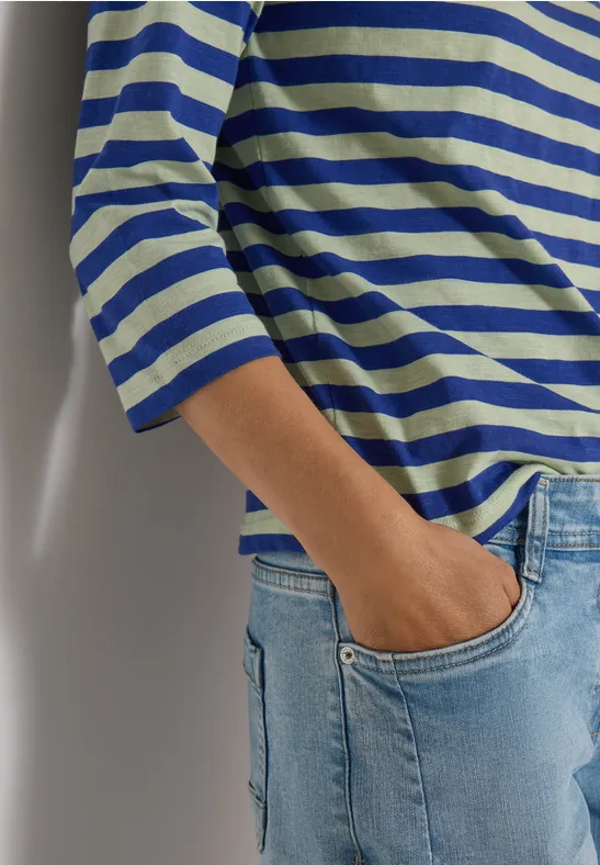 Persoon draagt blauw-wit gestreept 3/4-mouwshirt en spijkerbroek, hand in de zak.
