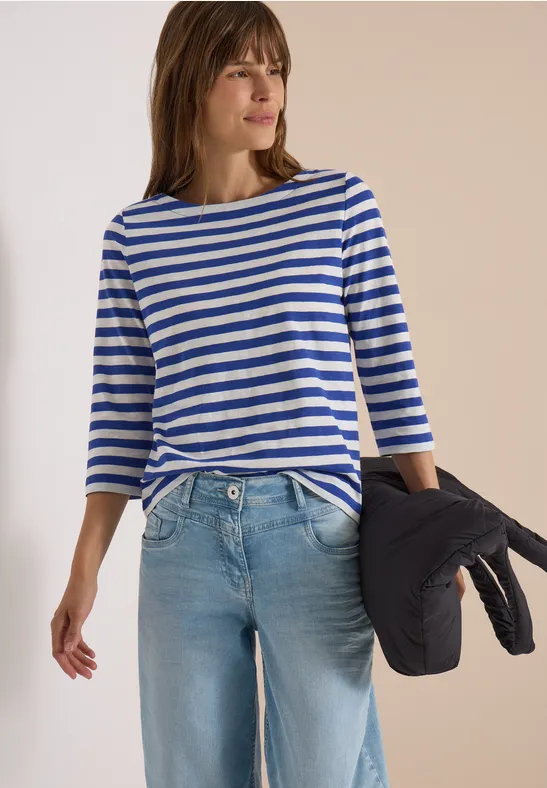 Vrouw in blauw-wit gestreept shirt, lichte spijkerbroek, met een donkere jas losjes over haar arm.