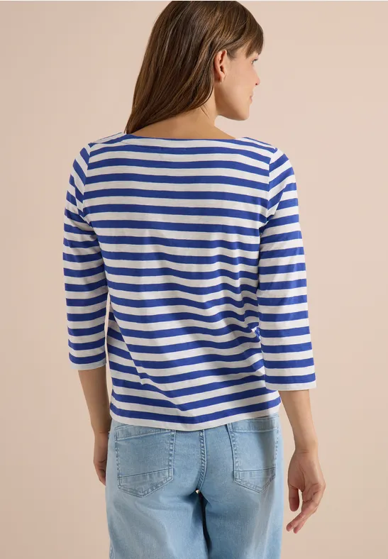 Vrouw van achteren in blauw-wit gestreept 3/4-mouwshirt en lichtblauwe spijkerbroek tegen beige achtergrond.