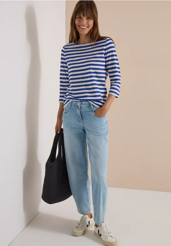 Vrouw lacht, draagt een blauw-wit gestreepte trui, lichte jeans, sneakers en zwarte tas voor een beige muur.