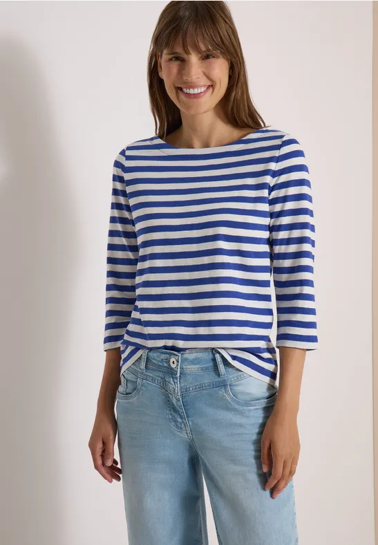 Vrouw glimlacht, draagt blauw-wit gestreept 3/4-mouw shirt en lichte spijkerbroek.