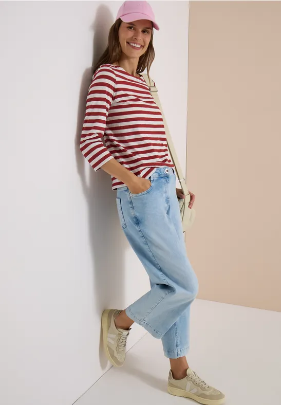 Vrouw met roze pet, rood-wit gestreept shirt, losse jeans en sneakers, glimlachend tegen een muur geleund.