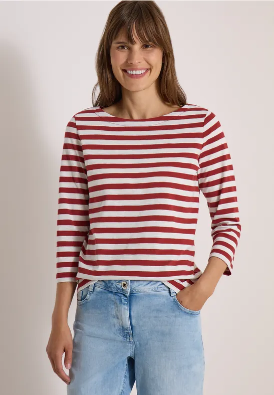 Een glimlachende vrouw draagt een rood-wit gestreept shirt en spijkerbroek tegen neutrale achtergrond.