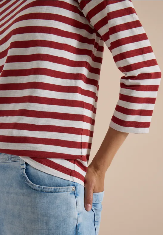 Persoon draagt een rood-wit gestreept 3/4-mouwen shirt en lichte jeans met hand in zak.