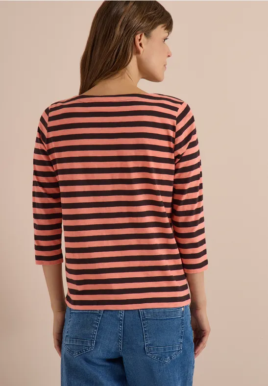 Vrouw van achter gezien met een roze-zwart gestreept 3/4-mouwshirt en blauwe spijkerbroek voor een beige muur.