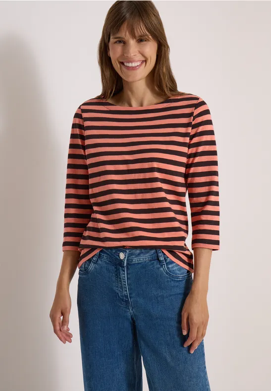 Lachende vrouw in roodzwart gestreept shirt en blauwe jeans voor een lichte achtergrond.