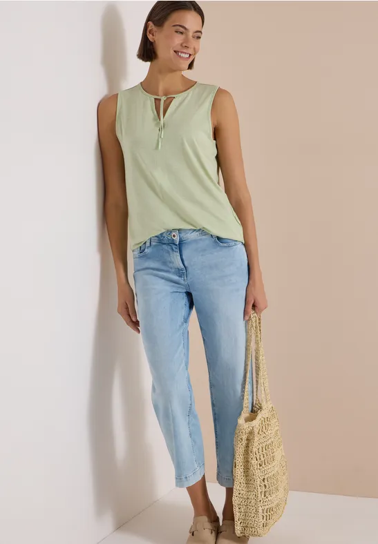 Frau in hellgrünem Top, hellblauen Jeans und Strohtasche lehnt lächelnd an heller Wand.