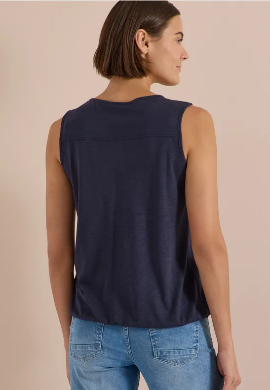 Eine Frau steht mit dem Rücken zur Kamera, trägt ein dunkelblaues ärmelloses Top und blaue Jeans vor beigem Hintergrund.