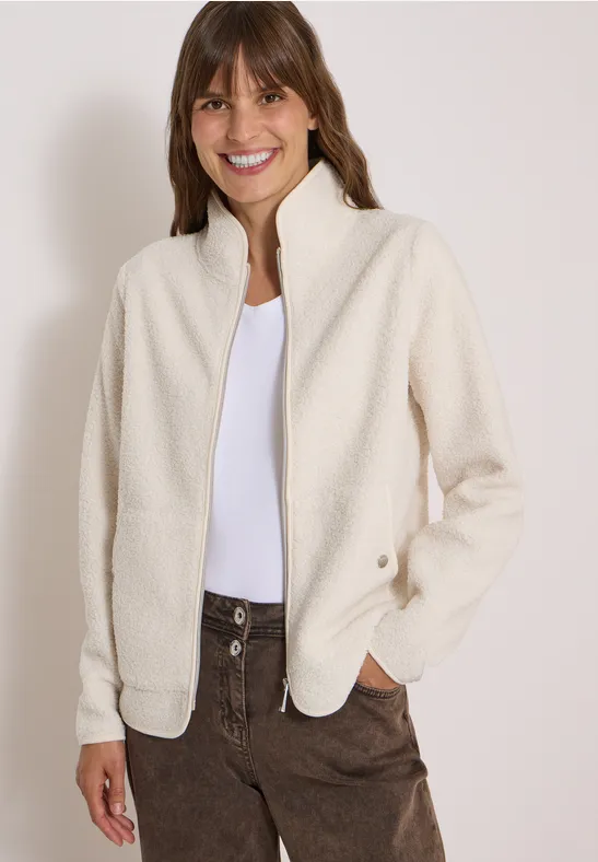 Frau in cremeweißer Fleecejacke, weißem Shirt und braunen Cordhosen, lächelt.