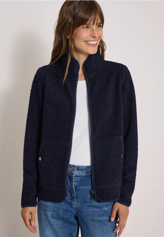 Frau trägt dunkelblaue Fleecejacke und Jeans, lächelt vor neutralem Hintergrund.
