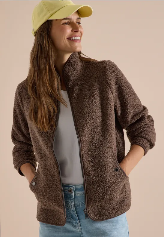 Lächelnde Frau in brauner Teddyfleece-Jacke, grauem Shirt und hellgrüner Cap.