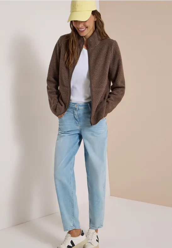 Frau in brauner Jacke, weißem T-Shirt, hellblauen Jeans, weißen Sneakern und beiger Cap.