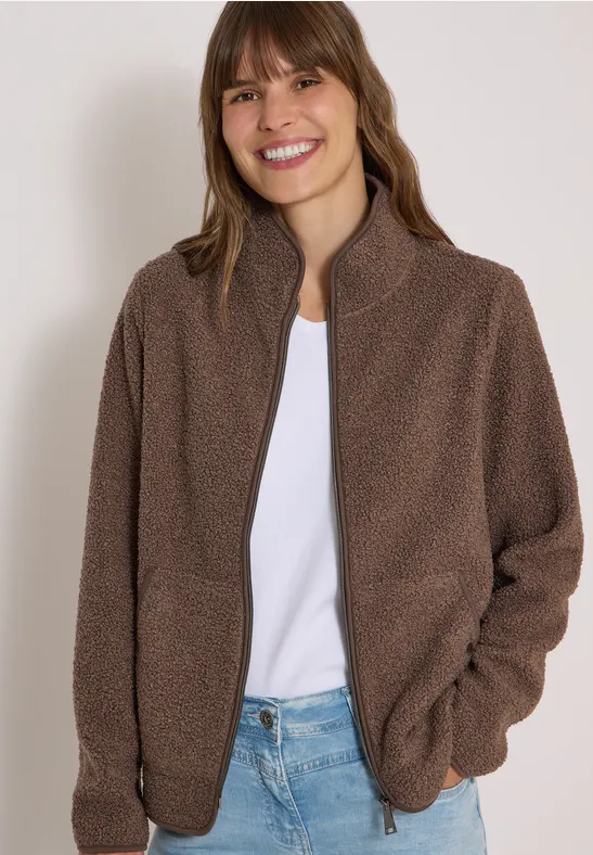 Frau lächelt in brauner Fleecejacke, weißem Shirt und Jeans vor neutralem Hintergrund