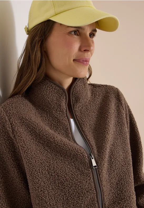 Frau mit gelber Kappe und brauner Fleecejacke, blickt seitlich.