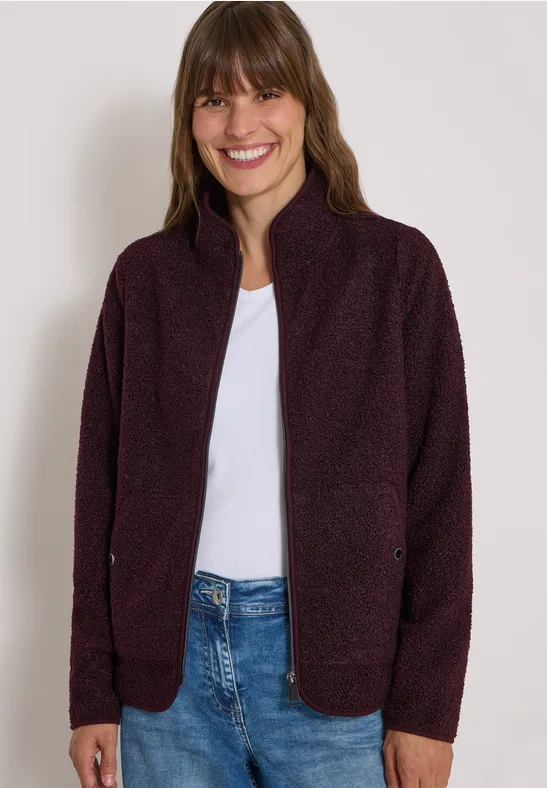 Lächelnde Frau trägt eine violette Strickjacke über weißem T-Shirt und Jeans.