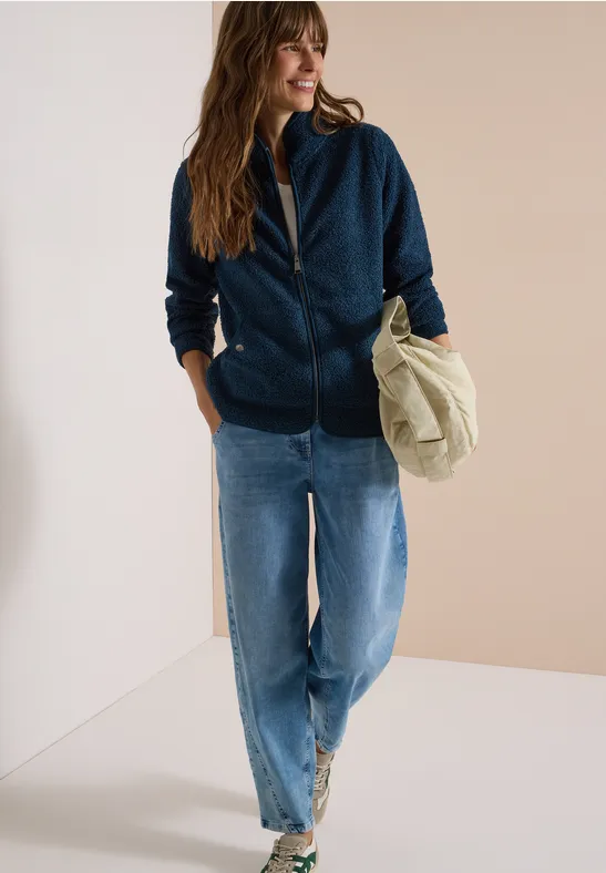 Frau in lässigem Outfit: dunkelblaue Kapuzenjacke, Jeans, weiße Sneaker, hält eine beige Tasche.