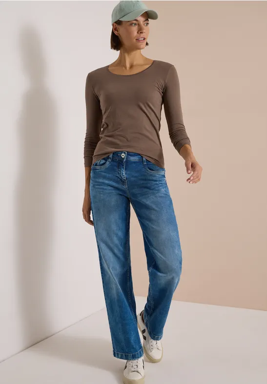 Une femme en casquette pose en jean bleu, haut marron et baskets blanches.